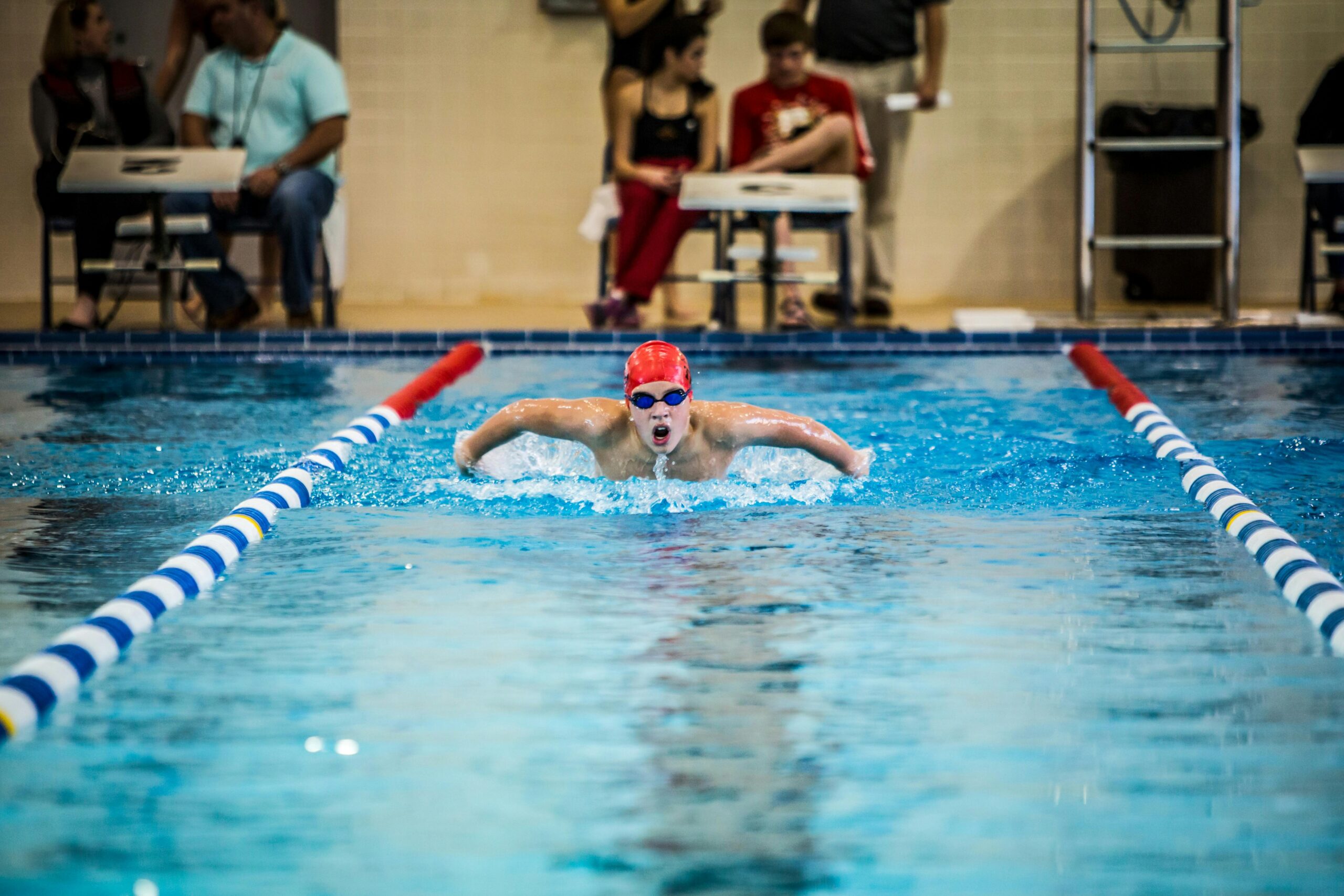 A young swimmer in a red cap competes in a butterfly stroke during an indoor pool race.