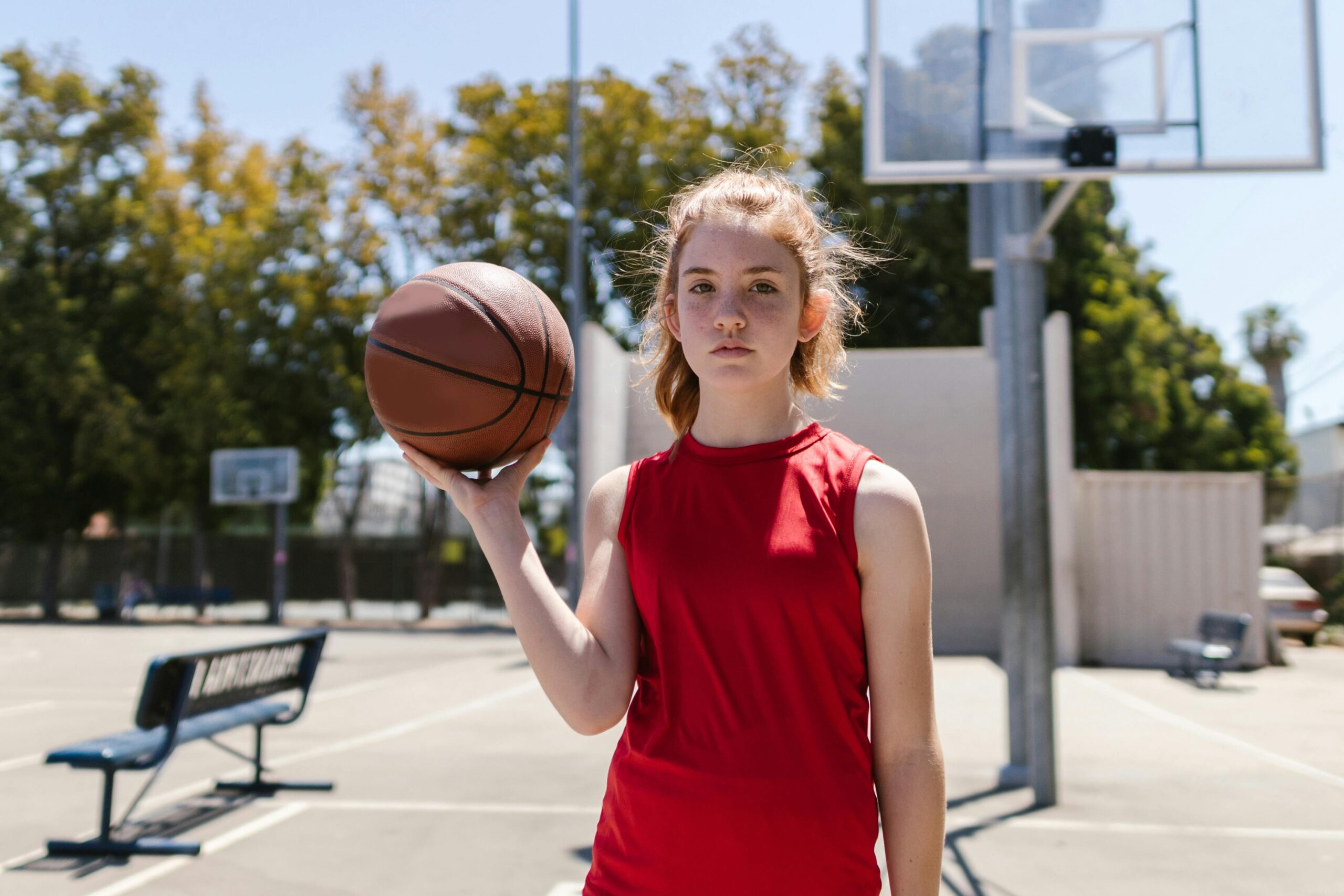Red-haired girl standing confidently with basketball on an outdoor court, ready to play.